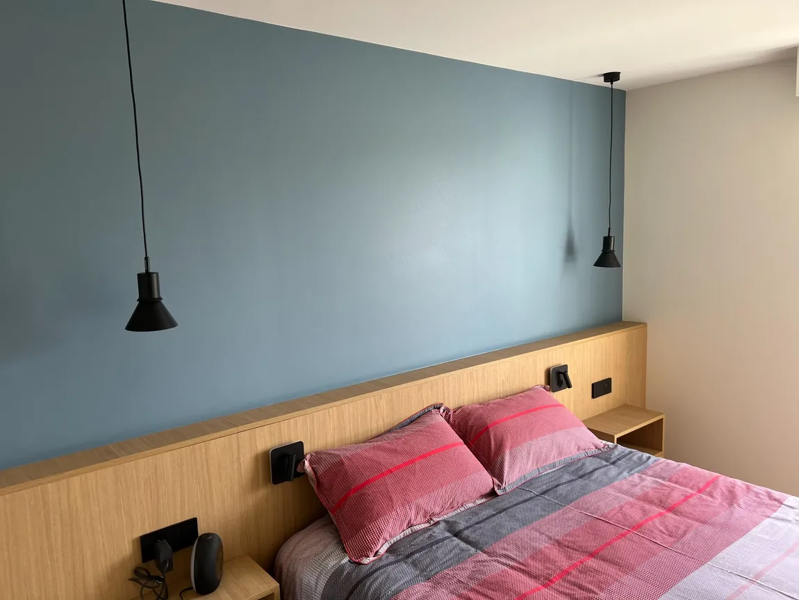 Tête de lit en bois sur mur bleu, avec suspensions noires, liseuses, prises et commandes intégrées.