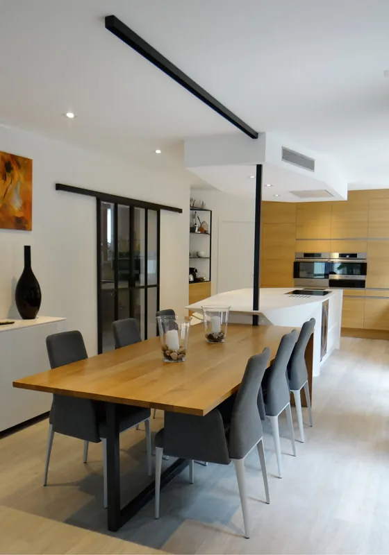 Cuisine avec îlot et grande table en bois ; une structure noire intègre le passage des alimentations.